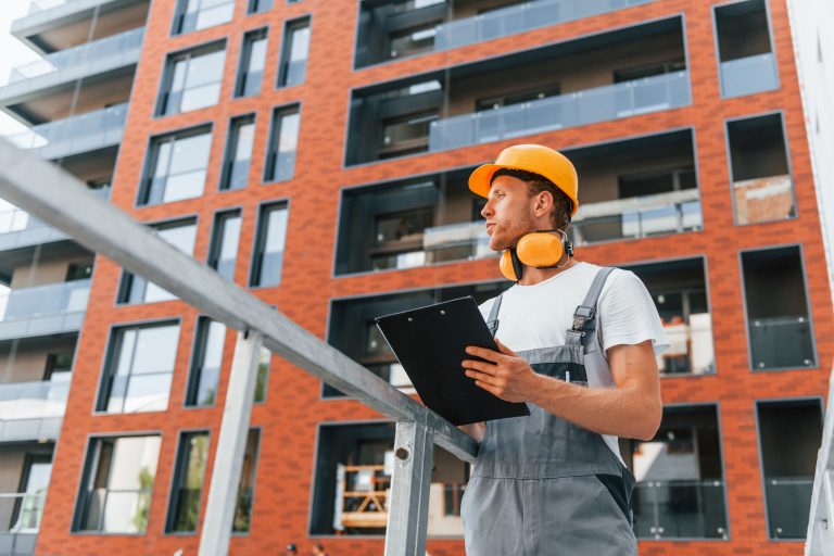 With documents in hands. Young man working in uniform at construction at daytime.